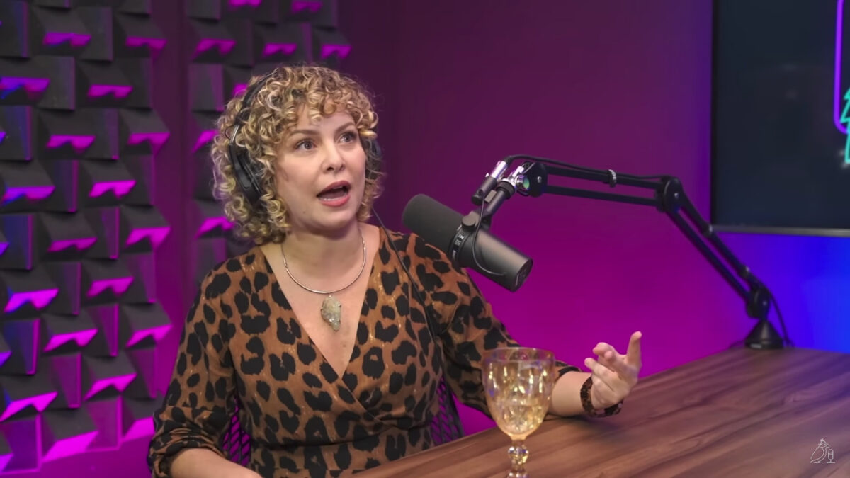Bárbara Borges de vestido animal print de onça, decotada, colar de prata sentada numa cadeira de ferro preta, com a mão na bancada do podcast Papagaio Falante
