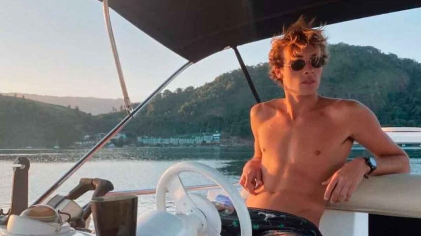 Bruno Krupp sem camisa e de óculos esculos, pilototando um pequeno barco em uma praia