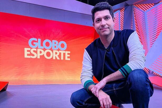 Caio Maciel agachado nos estúdios do Globo Esporte de SP, com o telão ao fundo, mostrando o logo do programa