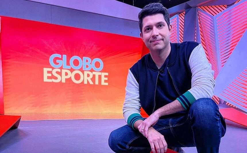 Caio Maciel agachado nos estúdios do Globo Esporte de SP, com o telão ao fundo, mostrando o logo do programa