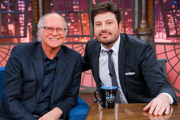 Paulo Roberto Galvão sorrindo, de palitó, camisa e calça jeans preta, com a mão no joelho, sentado num sofá azul ao lado de Danilo Gentili durante gravação do The Noite, do SBT