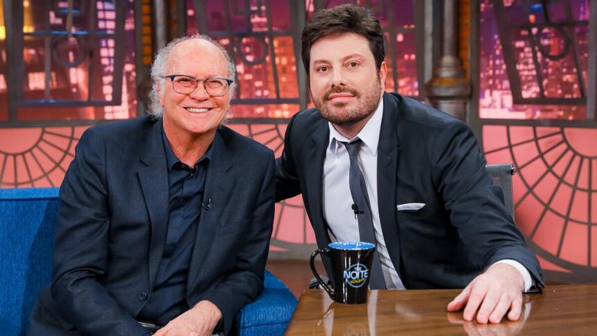 Paulo Roberto Galvão sorrindo, de palitó, camisa e calça jeans preta, com a mão no joelho, sentado num sofá azul ao lado de Danilo Gentili durante gravação do The Noite, do SBT