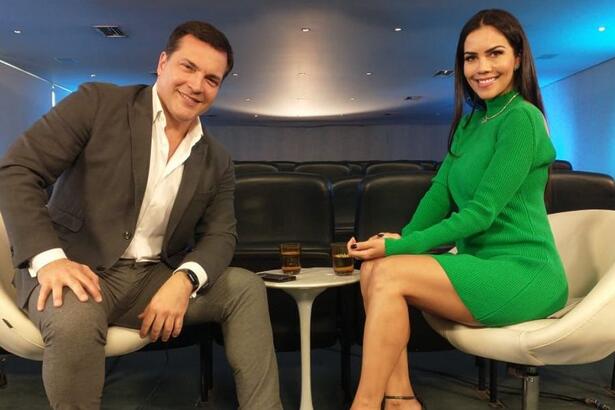 Daniel Boaventura de palitó preto, camisa branca e calça jeans cinza, sentado num banco branco ao lado de Daniela Albuquerque, de vestido verde, sorrindo, sentada num banco branco durante gravação de entrevista