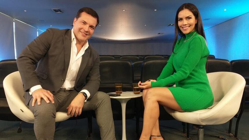 Daniel Boaventura de palitó preto, camisa branca e calça jeans cinza, sentado num banco branco ao lado de Daniela Albuquerque, de vestido verde, sorrindo, sentada num banco branco durante gravação de entrevista