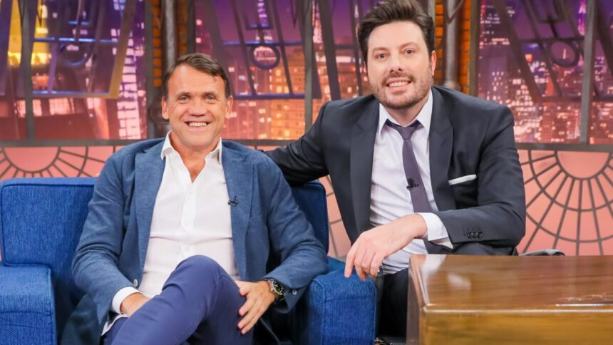 Dejan Petkovic de blazer e calça jeans azul e camisa social, com a perna cruzada e mão na coxa num sofá azul ao lado de Danilo Gentili durante gravação do The Noite, do SBT