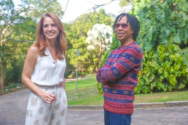 Poliana Brita de camisa branca e calça branca estampada sorrindo com Djavan, que está de camisa manga longa listrada em azul e rosa e calça jeans no jardim de sua casa durante gravação para o Fantástico da TV Globo