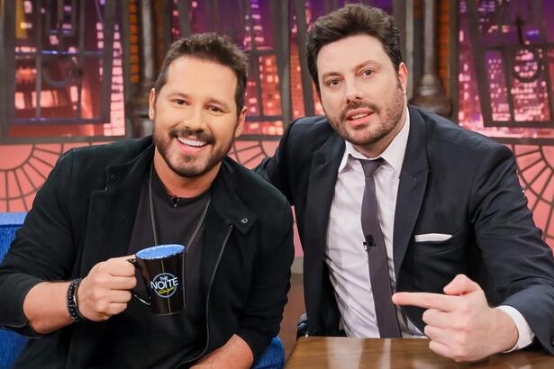 Dony de Nuccio de camisa e calça preta, sorrindo, segurando o caneco personalizado do The Noite, do SBT, ao lado do apresentador Danilo Gentili