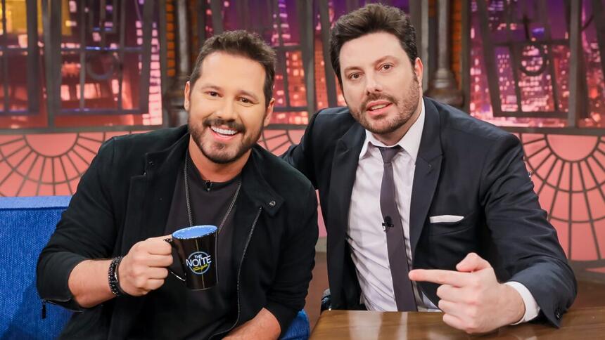 Dony de Nuccio de camisa e calça preta, sorrindo, segurando o caneco personalizado do The Noite, do SBT, ao lado do apresentador Danilo Gentili