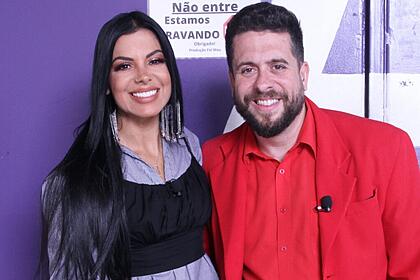 Evelyn Regly com top preto, camisa social roxa, sorrindo, cabelo preto solto ao lado de Mauricio Meireles, de blazer, camisa e calça vermelha, sorrindo, durante gravação do Foi Mau, da RedeTV!