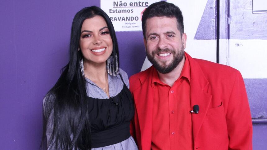 Evelyn Regly com top preto, camisa social roxa, sorrindo, cabelo preto solto ao lado de Mauricio Meireles, de blazer, camisa e calça vermelha, sorrindo, durante gravação do Foi Mau, da RedeTV!