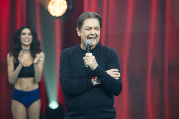 Fausto Silva sorrindo, de camisa manga longa preta, segurando o microfone com a mão esquerda durante gravação do seu programa na Band