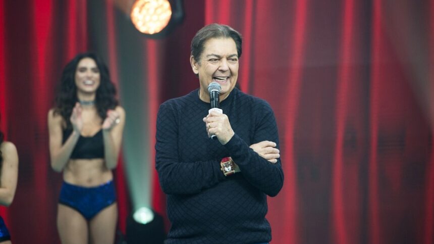 Fausto Silva sorrindo, de camisa manga longa preta, segurando o microfone com a mão esquerda durante gravação do seu programa na Band