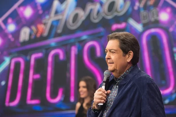 Faustão de camisa jeans azul escuro, de perfil, segurando microfone com a mão esquerda durante gravação do A Hora da Decisão, do Faustão na Band