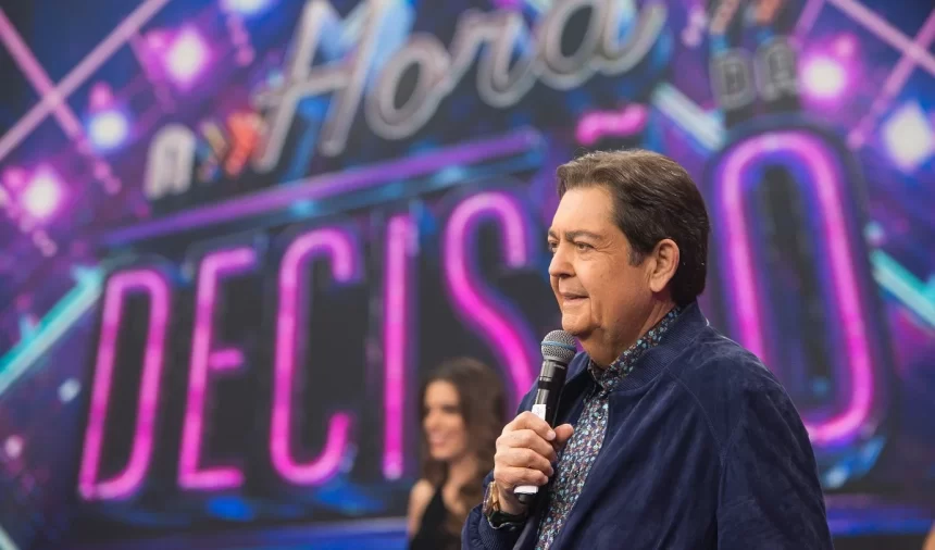 Faustão de camisa jeans azul escuro, de perfil, segurando microfone com a mão esquerda durante gravação do A Hora da Decisão, do Faustão na Band