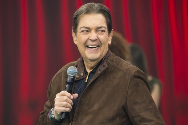 Fausto Silva sorrindo, de camisa manga longa marrom, segurando o microfone com a mão esquerda durante gravação do seu programa na Band