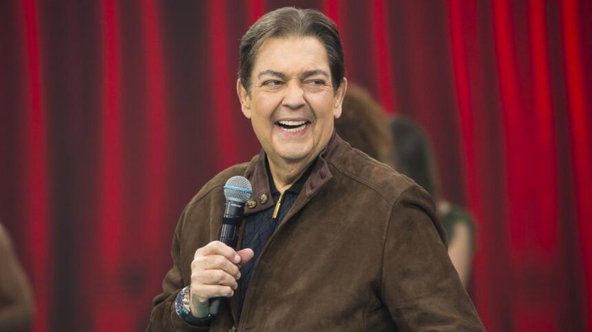 Fausto Silva sorrindo, de camisa manga longa marrom, segurando o microfone com a mão esquerda durante gravação do seu programa na Band