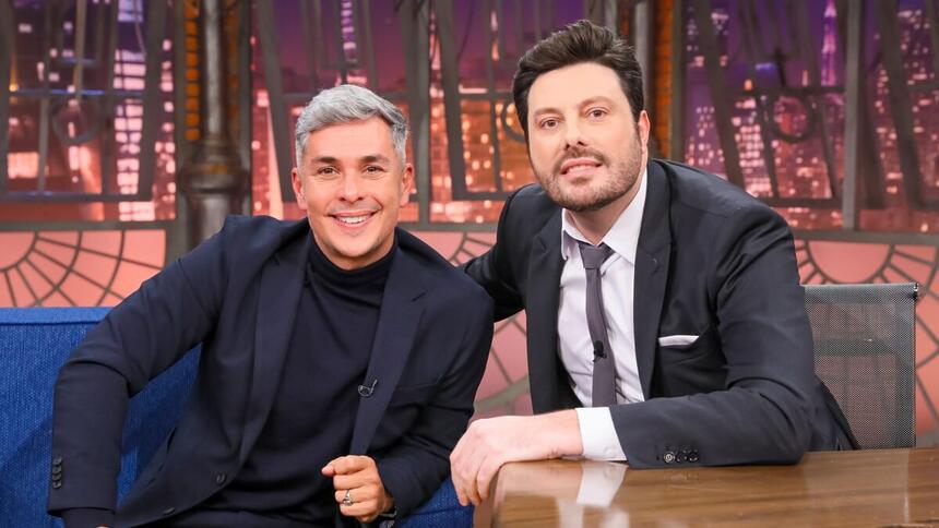 Ivan Moré de blazer e camisa preta, com o braço encostado no sofá azul, ao lado de Danilo Gentili durante gravação do The Noite, do SBT