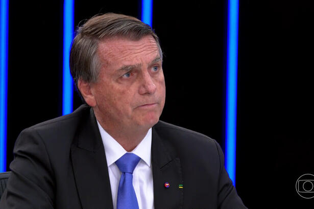 Jair Bolsonaro sério, de blazer preto e gravata azul, sentado na bancada do jornal Nacional durante debate