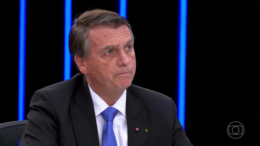 Jair Bolsonaro sério, de blazer preto e gravata azul, sentado na bancada do jornal Nacional durante debate