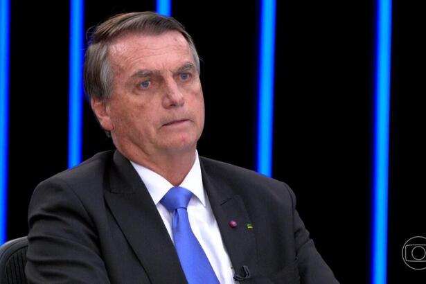 Jair Bolsonaro sério, de blazer preto e gravata azul, sentado na bancada do jornal Nacional durante debate