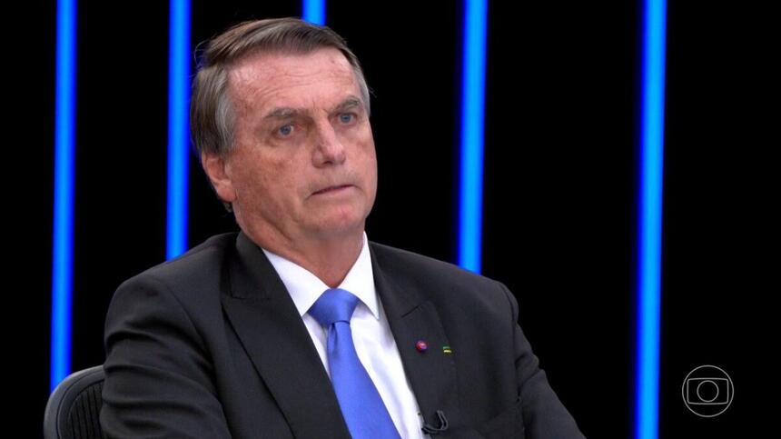 Jair Bolsonaro sério, de blazer preto e gravata azul, sentado na bancada do jornal Nacional durante debate