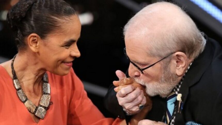 Marina Silva de colar, camisa vermelha, sorrindo, segurando a mão de Jô Soares durante gravação de programa de entrevistas na TV Globo