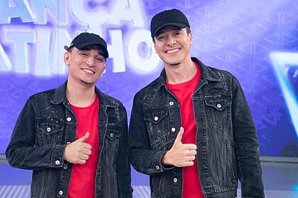 João Gomes e Rodrigo Faro com o mesmo chapéu preto, jaqueta jeans escura e camisa vermelha fazendo um legal com a mão direita durante gravação do Hora do Faro, da Record TV