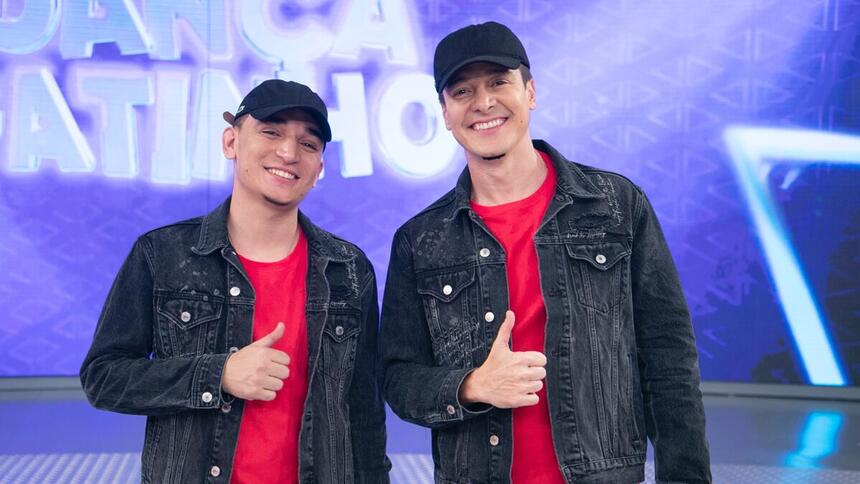 João Gomes e Rodrigo Faro com o mesmo chapéu preto, jaqueta jeans escura e camisa vermelha fazendo um legal com a mão direita durante gravação do Hora do Faro, da Record TV