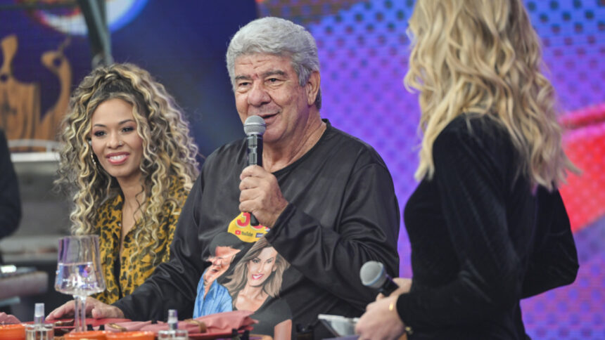 Juliana Diniz com body animal print, cabelo preso, sorrindo, ao lado de joel santana; joel Santana de camisa preta, segurando um microfone na mão esquerda, sentado durante gravação do Faustão na Band