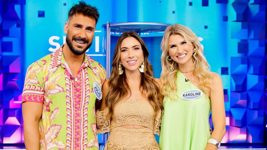 Júlio Rocha de camisa e bermuda estampada das cores vermelho, rosa e amarelo, sorrindo; Karoline Kleine de vestido verde claro, sorrindo; Patricia Abravanel de vestido bege, com brincos dourados, cabelo solto, sorrindo, durante gravação do Programa Silvio Santos, do SBT