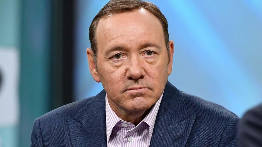 Kevin Spacey sério, com blazer azul e camisa manga longa roxa clara