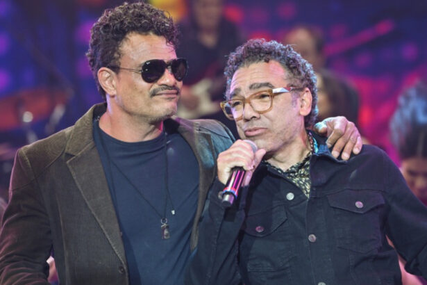 Leo Maia de óculos de sol preto, blazer preto e camisa azul marinho com a mão esquerna no ombro de Simoninha; Simoninha de camisa manga longa preta segurando o microfone preto com a mão direita durante gravação do Faustão na Band