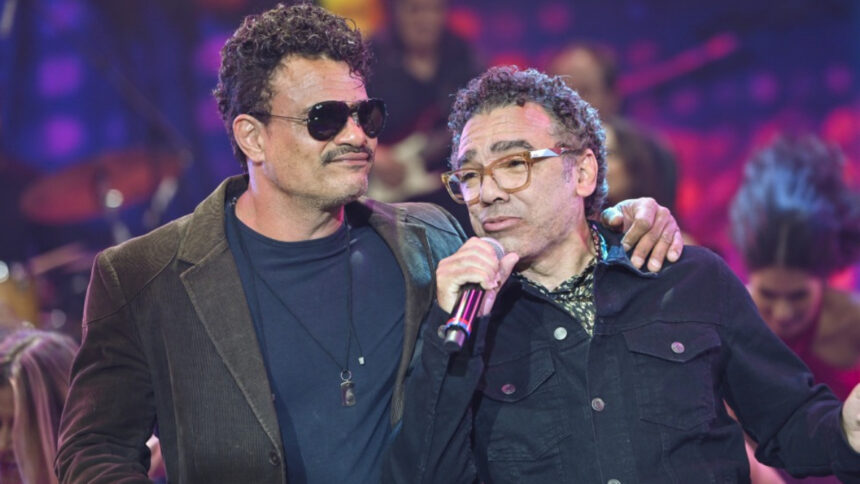 Leo Maia de óculos de sol preto, blazer preto e camisa azul marinho com a mão esquerna no ombro de Simoninha; Simoninha de camisa manga longa preta segurando o microfone preto com a mão direita durante gravação do Faustão na Band