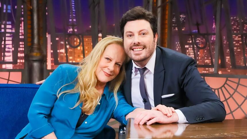 Lucinha Lins sorrindo, usando camisa azul e top preto, sentada no sofá azul, com a cabeça no ombro de Danilo Gentili e a mão na bancada do The Noite, do SBT