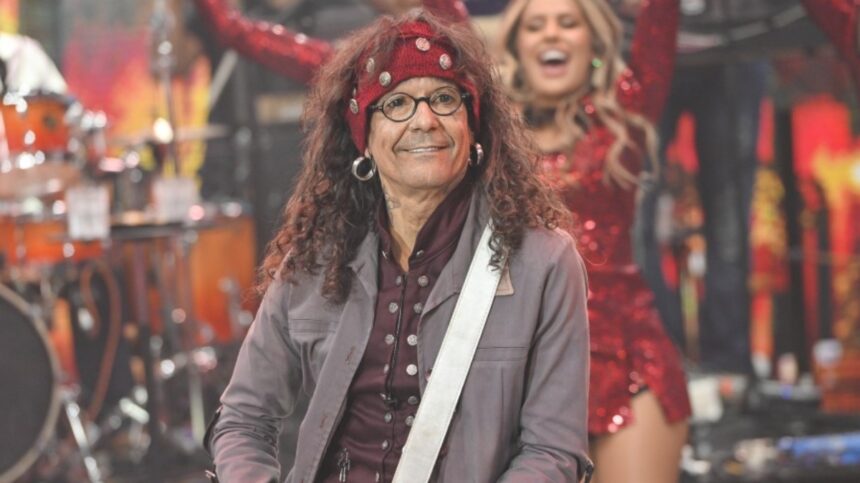 Cantor Luiz Caldas com blazer cinza, camisa preta e vermelha estampada e bandana na cabeça, tocando baixo, sorrindo durante performance do Faustão na Band