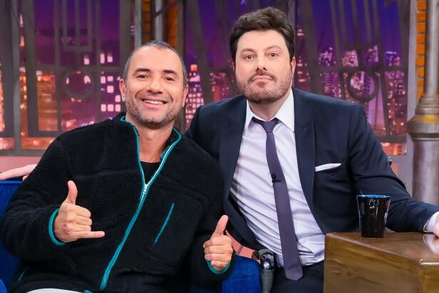 Marco Luque de camisa manga longa preta e flash verde, sorrindo, fazendo um legal e um hangloose com as mãos ao lado de Danilo Gentili