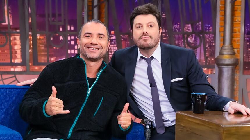 Marco Luque de camisa manga longa preta e flash verde, sorrindo, fazendo um legal e um hangloose com as mãos ao lado de Danilo Gentili