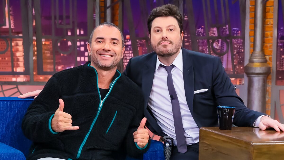 Marco Luque de camisa manga longa preta e flash verde, sorrindo, fazendo um legal e um hangloose com as mãos ao lado de Danilo Gentili