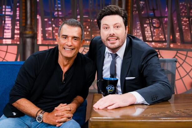 Marcos Pasquim sorrindo, de camisa preta e calça jeans, com as mãos na coxa, sentado no sofá azul ao lado de Danilo Gentili durante gravação do The Noite, do SBT