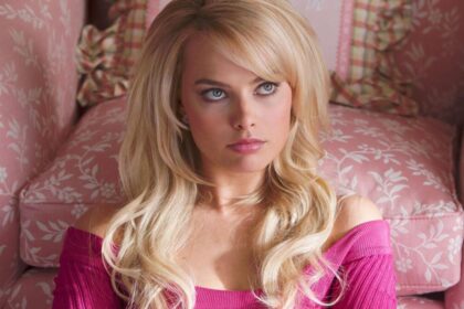 Margot Robbie caracterizada de Barbie de camisa rosa, peruca loira, sentada no chão, olhando pra cima, com semblante raivoso