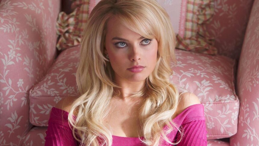 Margot Robbie caracterizada de Barbie de camisa rosa, peruca loira, sentada no chão, olhando pra cima, com semblante raivoso