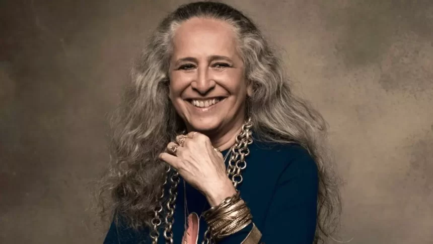 Maria Bethânia sorrindo, com a mão esquerda no queixo, cabelo solto, camisa azul escura, colares de ouro e prata durante ensaio fotografico