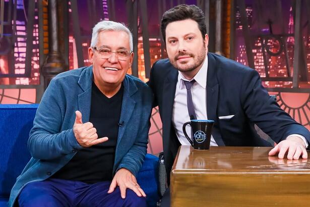 Mauro Naves de blazer azul, camisa preta, fazendo hangloose com a mão direita e a mão esquerda no joelho; Danilo Gentili de palitó e gravata preta sentado em sua bancada, sorrindo, durante gravação do The Noite, do SBT