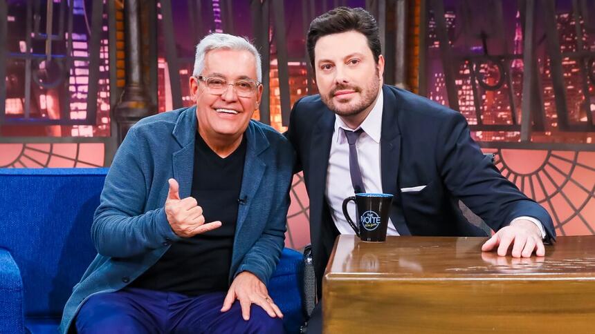 Mauro Naves de blazer azul, camisa preta, fazendo hangloose com a mão direita e a mão esquerda no joelho; Danilo Gentili de palitó e gravata preta sentado em sua bancada, sorrindo, durante gravação do The Noite, do SBT