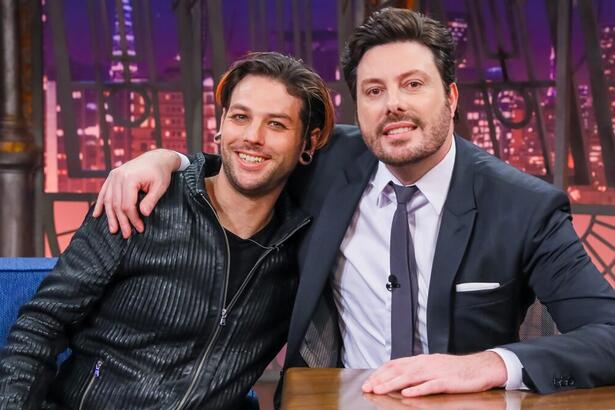 Navarone Garibaldi de camisa e calça preta, sorrindo, sentado no sofá azul ao lado de Danilo Gentili de palitó e gravata preta, sorrindo, com a mão esquerda na bancada durante gravação do The Noite, do SBT