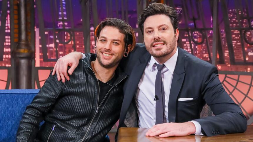 Navarone Garibaldi de camisa e calça preta, sorrindo, sentado no sofá azul ao lado de Danilo Gentili de palitó e gravata preta, sorrindo, com a mão esquerda na bancada durante gravação do The Noite, do SBT