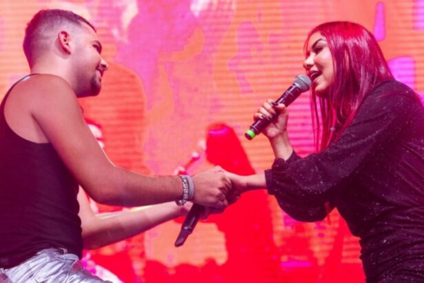 Pedro Sampaio de camisa regata preta e calça jeans segurando a mão de Priscila Sena, que está de vestido preto, segurando o microfone com a mão direita durante apresentação na Arena Pernambuco