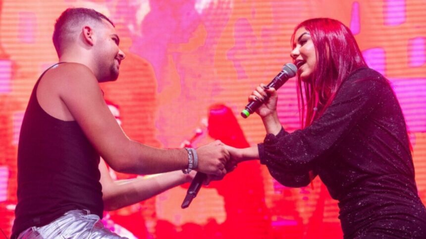 Pedro Sampaio de camisa regata preta e calça jeans segurando a mão de Priscila Sena, que está de vestido preto, segurando o microfone com a mão direita durante apresentação na Arena Pernambuco