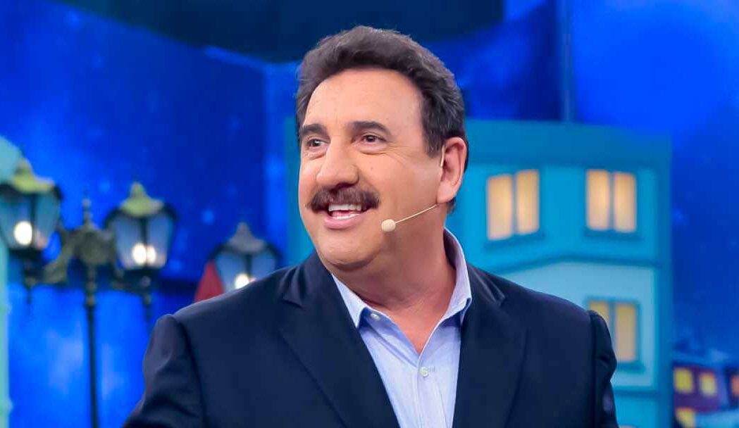 Ratinho de blazer azul escuro e camisa social azul claro, sorrindo, de perfil, microfone no rosto durante gravação de seu programa no SBT