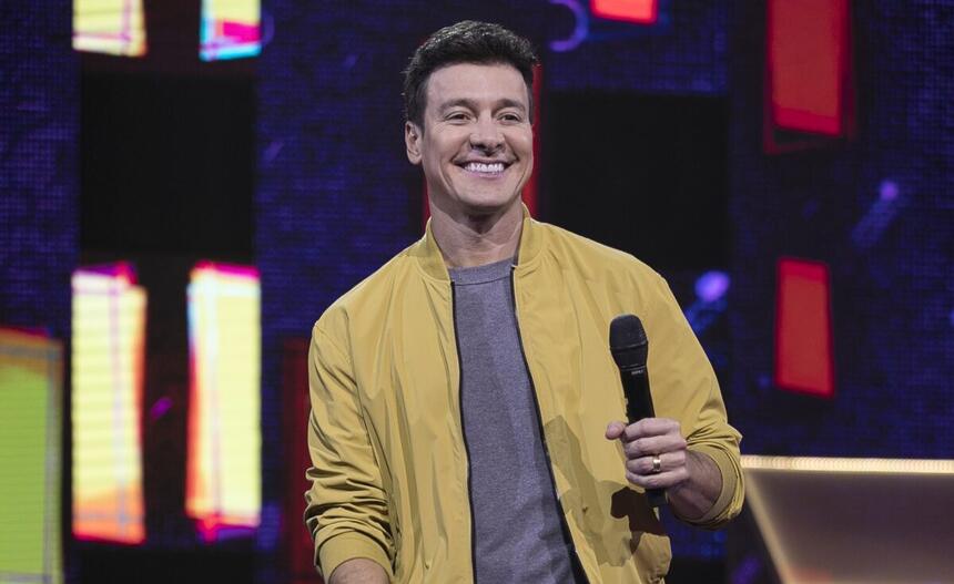 Rodrigo Faro segurando um microfone e sorridente, nos estúdios do Canta Comigo Teen